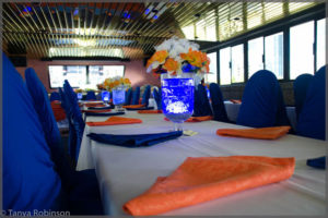 Floral centrepiece with white and orange flowers. Blue and orange napkins with blue chair covers.