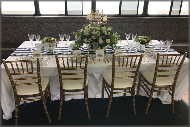 Table set up for Nautical decor dinner cruise with white roses and gold chairs.